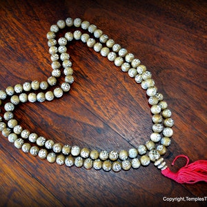 May include: A beaded mala necklace with a red tassel, resting on a dark wooden surface. The beads are light brown with white markings. The necklace is arranged in a heart shape.