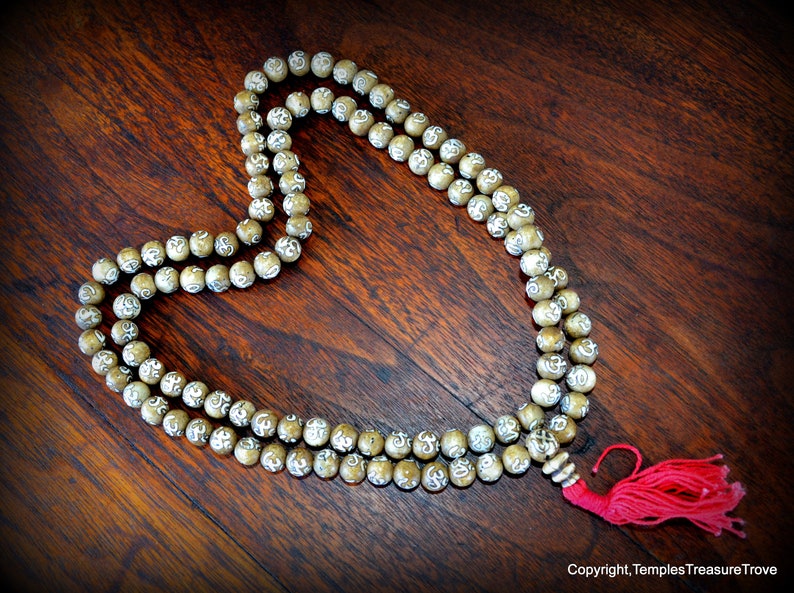 May include: A beaded mala necklace with a red tassel, resting on a dark wooden surface. The beads are light brown with white markings. The necklace is arranged in a heart shape.