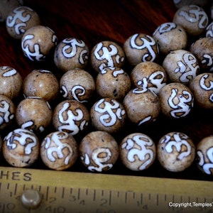 May include: Close-up of a collection of round, light brown beads with white, stylized markings. The beads are arranged on a dark wooden surface next to a ruler marked in inches. The beads appear to be approximately 0.75 inches in diameter.