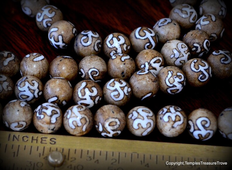 May include: Close-up of a collection of round, light brown beads with white, stylized markings. The beads are arranged on a dark wooden surface next to a ruler marked in inches. The beads appear to be approximately 0.75 inches in diameter.