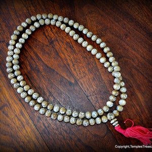 May include: A beaded mala necklace with light brown beads, each etched with a white symbol. The necklace has a red tassel and is displayed on a dark wooden surface. The beads are strung together.