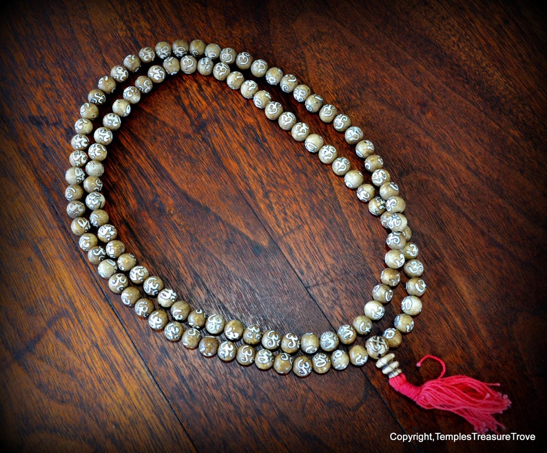 May include: A beaded mala necklace with light brown beads, each etched with a white symbol. The necklace has a red tassel and is displayed on a dark wooden surface. The beads are strung together.