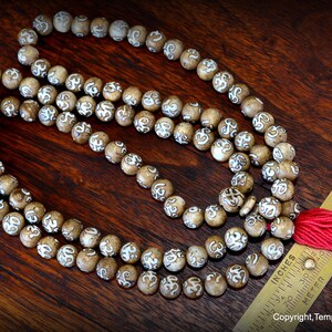 May include: A strand of prayer beads with light brown, round beads, each etched with a white design. A red tassel hangs from the end. A ruler with inches and centimeters is next to the beads. The beads are on a dark wood surface.