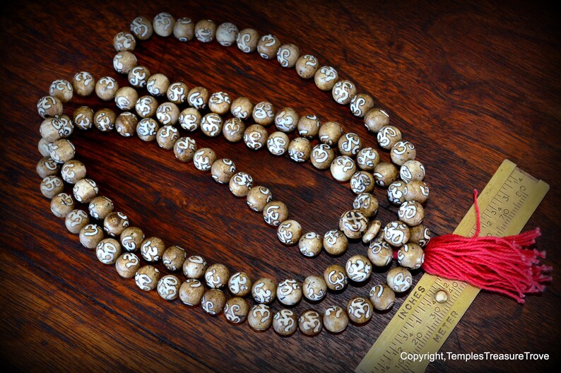 May include: A strand of prayer beads with light brown, round beads, each etched with a white design. A red tassel hangs from the end. A ruler with inches and centimeters is next to the beads. The beads are on a dark wood surface.
