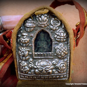 May include: A silver-colored religious pendant with intricate embossed designs and a small figure inside a central arch. The pendant is encased in a tan fabric pouch with a red and gold strap. The image is from Copyright TemplesTreasureTrove.