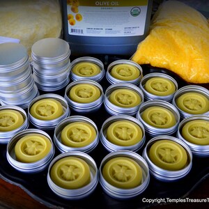 May include: A tray of small round metal tins filled with yellow beeswax with a bee design. The tins are arranged in a grid pattern on a black tray.