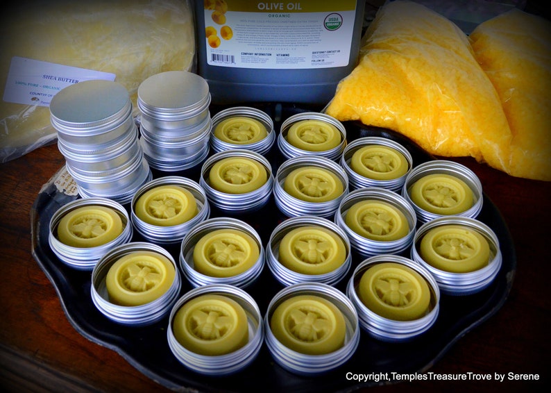 May include: A tray of small round metal tins filled with yellow beeswax with a bee design. The tins are arranged in a grid pattern on a black tray.