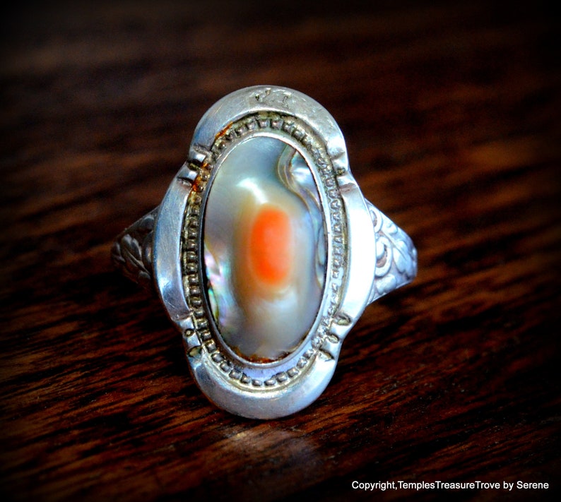 May include: A silver ring with an oval-shaped abalone shell inlay. The shell has a light orange center and iridescent colors.
