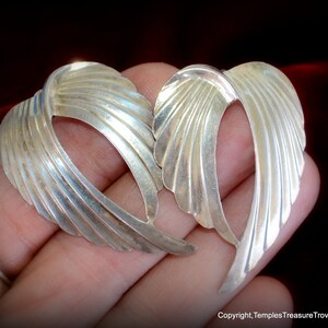 May include: A pair of silver earrings shaped like wings. The earrings are made of smooth, textured metal and have a delicate, feminine design.