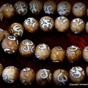 May include: Close-up of a strand of light brown beads with white, stylized symbols. The beads are round and evenly spaced, with a glossy finish. The background is a dark, wooden surface.