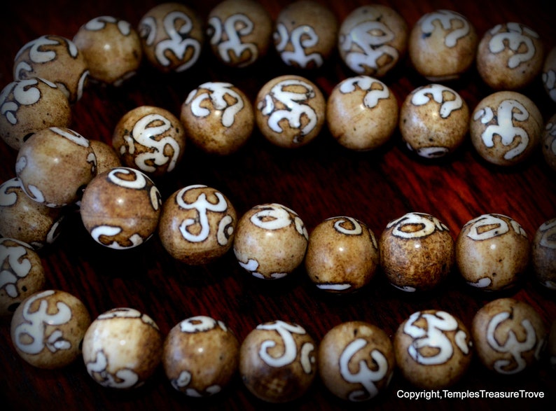 May include: Close-up of a strand of light brown beads with white, stylized symbols. The beads are round and evenly spaced, with a glossy finish. The background is a dark, wooden surface.