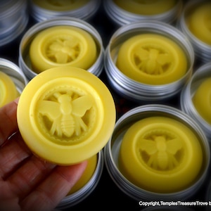 May include: Multiple small round containers of yellow beeswax balm with a bee design embossed on the top. The balm is in a silver tin.