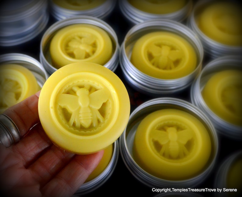 May include: Multiple small round containers of yellow beeswax balm with a bee design embossed on the top. The balm is in a silver tin.