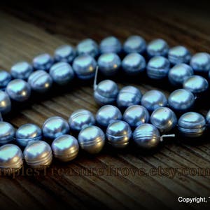 May include: A strand of blue freshwater pearls, with a slight iridescent sheen, is displayed on a wooden surface. The pearls are round and smooth, and they are arranged in a loose circle.