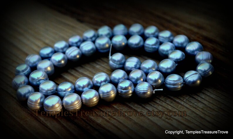May include: A strand of blue freshwater pearls, with a slight iridescent sheen, is displayed on a wooden surface. The pearls are round and smooth, and they are arranged in a loose circle.