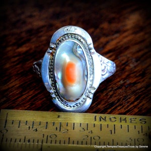 May include: A silver ring with an oval-shaped abalone shell inlay. The shell has a light orange center and iridescent colors. The ring has a decorative design around the shell.