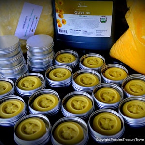May include: A close-up of 17 small round metal tins filled with a yellow substance. The tins are arranged in rows on a black surface. The tins have a bee design on the top.