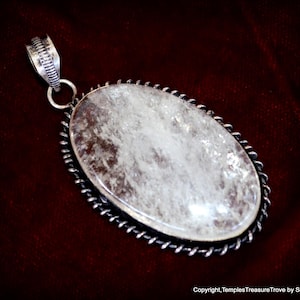 May include: A silver pendant with a large, oval-shaped clear quartz crystal. The crystal has a cloudy, white appearance and is set in a silver bezel with a twisted rope design.
