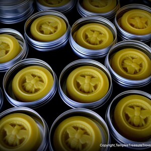 May include: A collection of small, round, silver tins with lids, each containing a yellow, bee-shaped wax balm. The tins are arranged in rows on a dark surface.