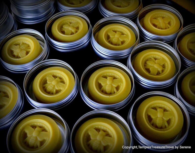 May include: A collection of small, round, silver tins with lids, each containing a yellow, bee-shaped wax balm. The tins are arranged in rows on a dark surface.