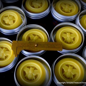May include: A close-up of several small round metal tins filled with a yellow beeswax balm. Each tin has a bee design embossed on the top. The tins are arranged in a grid pattern on a dark surface.