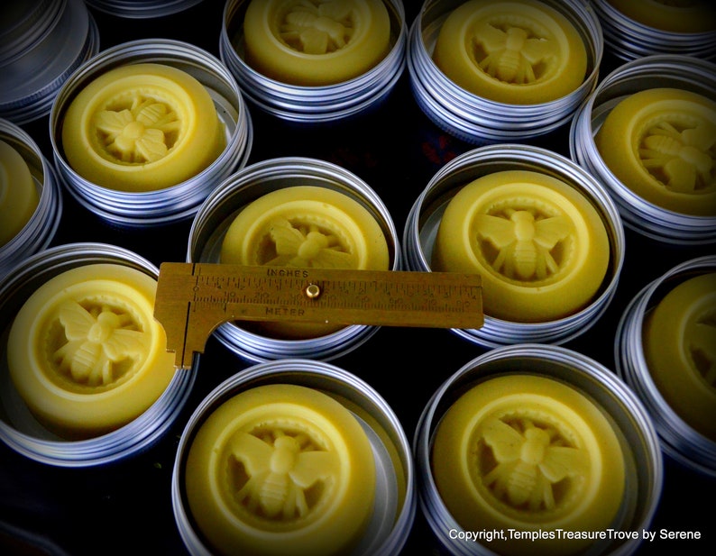 May include: A close-up of several small round metal tins filled with a yellow beeswax balm. Each tin has a bee design embossed on the top. The tins are arranged in a grid pattern on a dark surface.