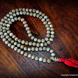 May include: A beige mala bead necklace with white etched symbols, a red tassel, and a silver-colored spacer. The beads are arranged on a dark wooden surface. The necklace is a spiritual or meditation accessory.