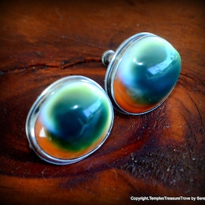 May include: A pair of oval stud earrings. Each earring has a polished, domed stone with a gradient of green, orange, and blue. The stones are set in a silver-coloured metal bezel. The earrings are displayed on a dark wooden surface.