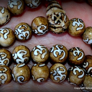 May include: Close-up of a strand of prayer beads. The beads are round, with a light brown base color and white, stylized symbols. The beads are strung together on a red cord. The image is well-lit, highlighting the details of the beads.
