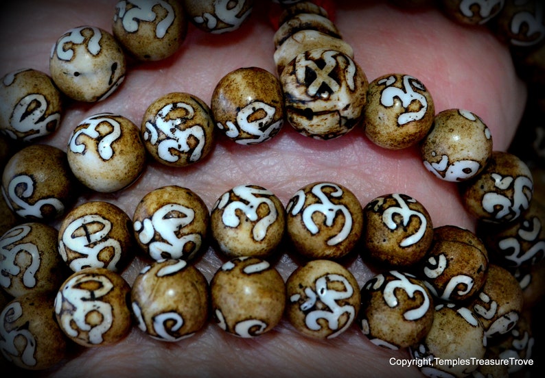 May include: Close-up of a strand of prayer beads. The beads are round, with a light brown base color and white, stylized symbols. The beads are strung together on a red cord. The image is well-lit, highlighting the details of the beads.