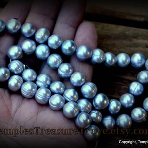 May include: A strand of light blue freshwater pearls, strung on a white cord, with a silver clasp. The pearls are round and smooth, and they are arranged in a single row. The pearls are displayed on a hand, with the background out of focus.