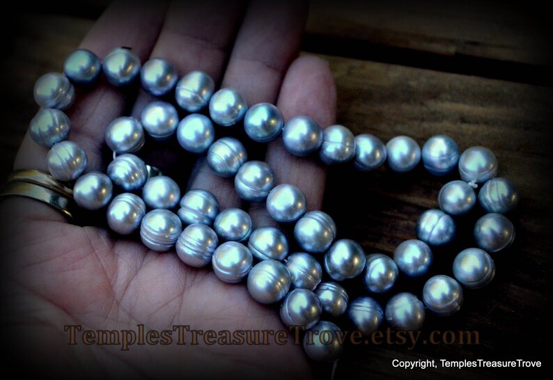 May include: A strand of light blue freshwater pearls, strung on a white cord, with a silver clasp. The pearls are round and smooth, and they are arranged in a single row. The pearls are displayed on a hand, with the background out of focus.