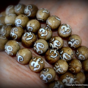 May include: Close-up of a strand of prayer beads. The beads are round, light brown, and feature a white Om symbol. The beads are held in a hand, with the focus on the beads themselves. The beads are likely used for meditation.