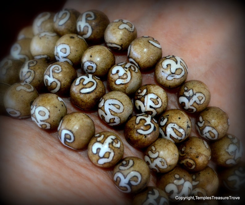 May include: Close-up of a strand of prayer beads. The beads are round, light brown, and feature a white Om symbol. The beads are held in a hand, with the focus on the beads themselves. The beads are likely used for meditation.