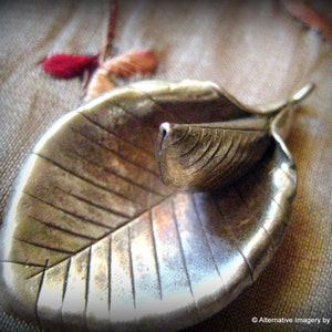 May include: A silver leaf pendant with a detailed, textured design. The leaf is folded in half, with the top half forming a smaller leaf shape.