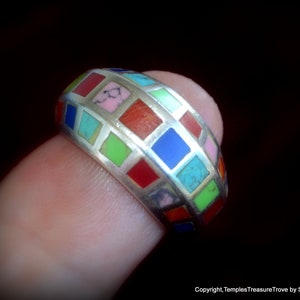 Peut inclure: Une bague en argent avec un motif en mosaïque. La bague présente une série de carrés colorés en rouge, bleu, vert, rose et turquoise. La bague est tenue par un doigt sur un fond sombre.