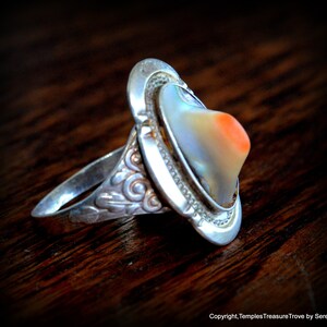 May include: A silver ring with an abalone shell inlay. The shell has a pearlescent sheen with a hint of orange. The ring band is decorated with a floral design.