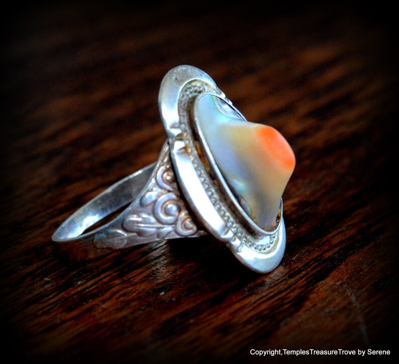 May include: A silver ring with an abalone shell inlay. The shell has a pearlescent sheen with a hint of orange. The ring band is decorated with a floral design.
