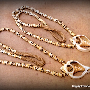 Puede incluir: Dos collares de hueso blanco con cuentas de calavera y colgantes de concha. Los collares están sobre un fondo de tela marrón.