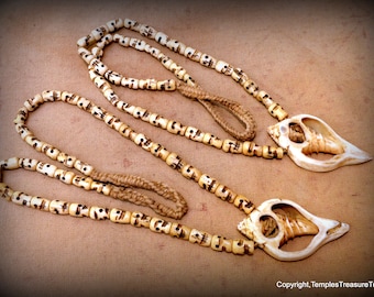 May include: Two white bone necklaces with skull beads and shell pendants. The necklaces are on a brown fabric background.