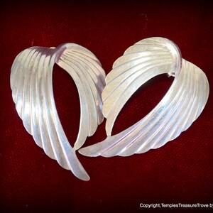May include: A pair of silver earrings with a unique, abstract design. The earrings are shaped like a wing or a leaf, with a series of curved lines that create a flowing, organic look.