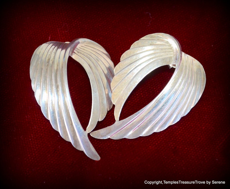 May include: A pair of silver earrings with a unique, abstract design. The earrings are shaped like a wing or a leaf, with a series of curved lines that create a flowing, organic look.