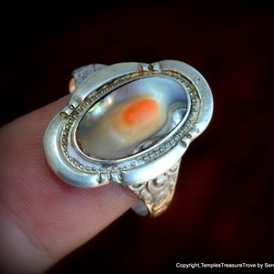 May include: A silver ring with an oval-shaped abalone shell inlay. The shell has a pearlescent white and iridescent blue and green color with a small orange spot in the center.