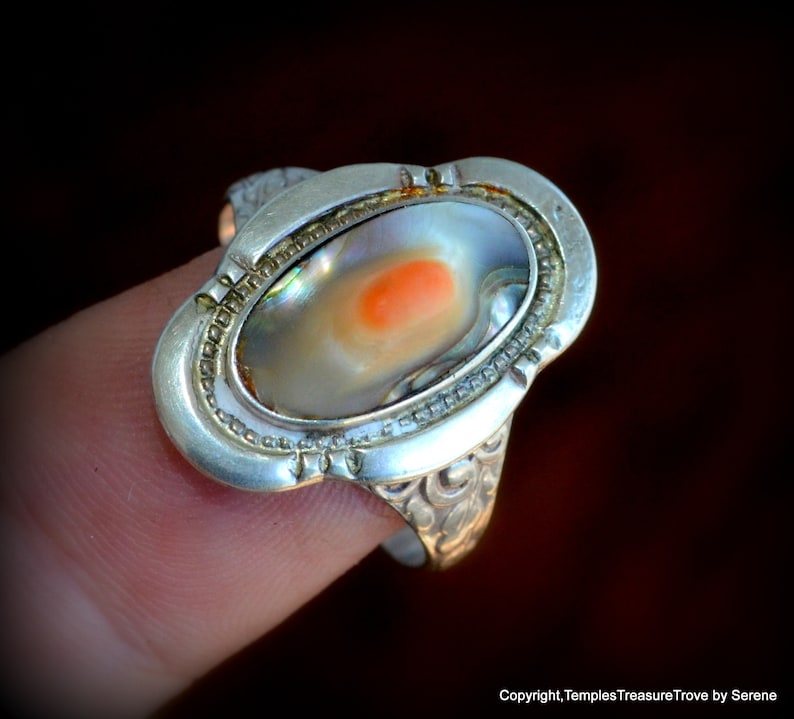 May include: A silver ring with an oval-shaped abalone shell inlay. The shell has a pearlescent white and iridescent blue and green color with a small orange spot in the center.
