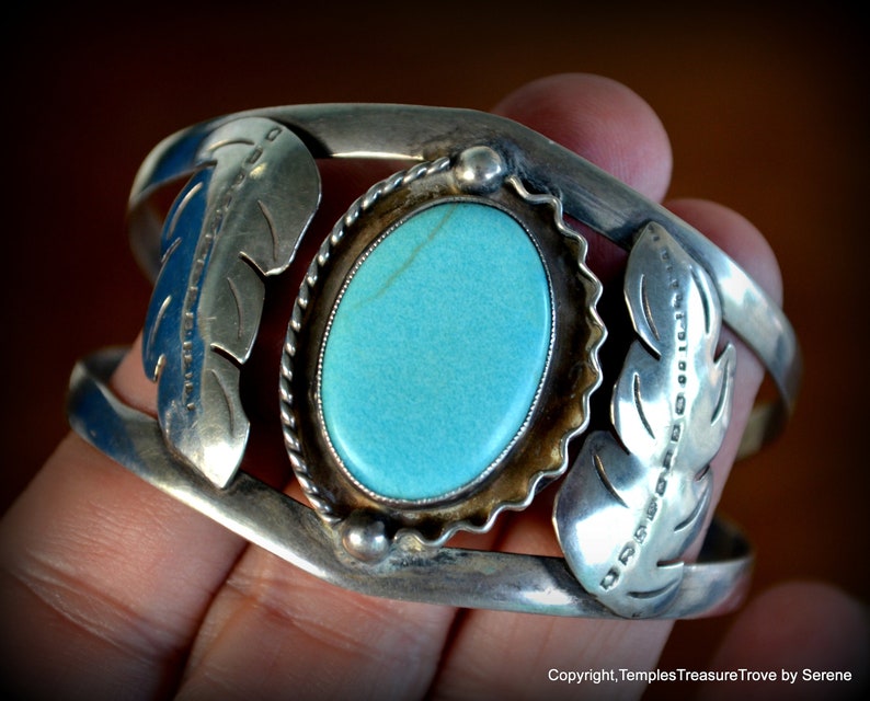 Puede incluir: Un brazalete de plata con una piedra turquesa engastada en un bisel de plata. El brazalete est&aacute; decorado con dos dise&ntilde;os de plumas de plata a cada lado de la piedra.