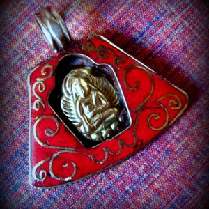 May include: A red and gold pendant with a detailed design. The pendant features a small, gold, seated figure in the center.
