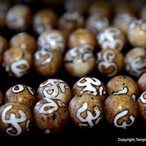 May include: Close-up of numerous brown beads with white, stylized symbols. The beads have a natural, organic appearance, with variations in color and texture. The symbols are painted in a contrasting white, creating a visual focal point. The beads are likely used for jewelry or decorative purposes.