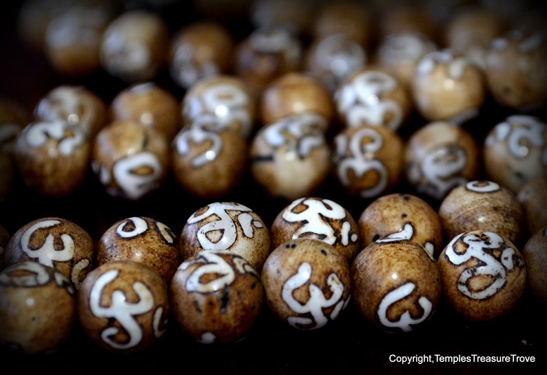 May include: Close-up of numerous brown beads with white, stylized symbols. The beads have a natural, organic appearance, with variations in color and texture. The symbols are painted in a contrasting white, creating a visual focal point. The beads are likely used for jewelry or decorative purposes.