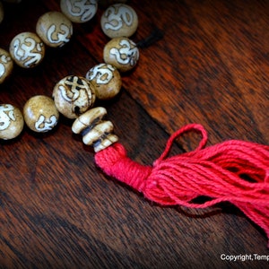 May include: Close-up of a beaded necklace with tan-colored beads featuring white symbols. The necklace has a red tassel and is displayed on a dark wooden surface. The beads are approximately 0.75 inches in diameter.