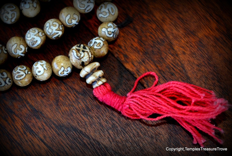 May include: Close-up of a beaded necklace with tan-colored beads featuring white symbols. The necklace has a red tassel and is displayed on a dark wooden surface. The beads are approximately 0.75 inches in diameter.